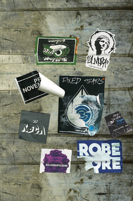 Various stickers are placed on a wooden surface, including designs with text such as 'Fixed Stars,' 'Bolognastickers,' and 'Tattoo Studio.' Some stickers show wear and tear, with edges peeling or faded areas. The background consists of rustic wooden planks with visible grain and weathering.