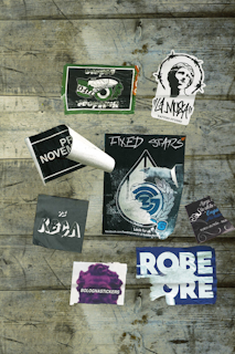 Various stickers are placed on a wooden surface, including designs with text such as 'Fixed Stars,' 'Bolognastickers,' and 'Tattoo Studio.' Some stickers show wear and tear, with edges peeling or faded areas. The background consists of rustic wooden planks with visible grain and weathering.