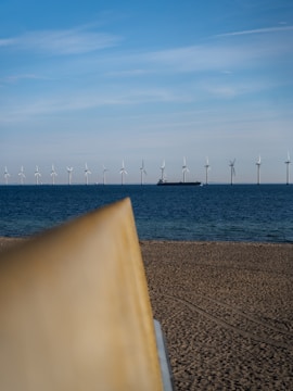Wind turbines are lined up in the distance across the sea, with a large vessel visible among them. In the foreground, a sandy beach is partially visible with a blurred object, possibly a part of a surfboard or similar item, also in view. The sky is clear with minimal clouds.