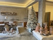 A rustic interior space featuring pottery and ceramic crafts displayed on tables and shelves. A frosted, decorated Christmas tree stands in the corner, adding a festive touch. The walls are textured concrete, complementing the earthy tones of the pottery. Pendant lighting and wooden furniture contribute to a cozy, artisan atmosphere.