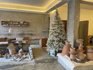 A rustic interior space featuring pottery and ceramic crafts displayed on tables and shelves. A frosted, decorated Christmas tree stands in the corner, adding a festive touch. The walls are textured concrete, complementing the earthy tones of the pottery. Pendant lighting and wooden furniture contribute to a cozy, artisan atmosphere.