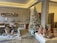A rustic interior space featuring pottery and ceramic crafts displayed on tables and shelves. A frosted, decorated Christmas tree stands in the corner, adding a festive touch. The walls are textured concrete, complementing the earthy tones of the pottery. Pendant lighting and wooden furniture contribute to a cozy, artisan atmosphere.