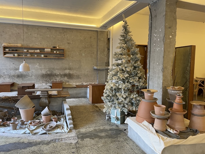 A rustic interior space featuring pottery and ceramic crafts displayed on tables and shelves. A frosted, decorated Christmas tree stands in the corner, adding a festive touch. The walls are textured concrete, complementing the earthy tones of the pottery. Pendant lighting and wooden furniture contribute to a cozy, artisan atmosphere.