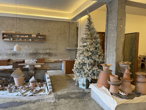 A rustic interior space featuring pottery and ceramic crafts displayed on tables and shelves. A frosted, decorated Christmas tree stands in the corner, adding a festive touch. The walls are textured concrete, complementing the earthy tones of the pottery. Pendant lighting and wooden furniture contribute to a cozy, artisan atmosphere.