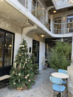 An outdoor courtyard features a well-decorated Christmas tree adorned with gold ornaments and candles. The scene includes a small round table with two blue chairs, set against a backdrop of brick walls and a wrought iron balcony. There are plants around, adding greenery to the urban setting.