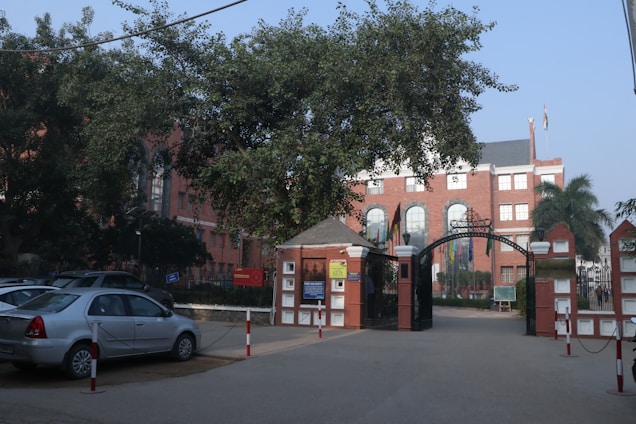 A large brick building is visible through a wrought iron gate, surrounded by various trees and parked cars. The building has several tall, arched windows and multiple flags at the entrance. The overall setting suggests a formal or institutional location, such as a school or university.