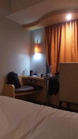 A cozy hotel room featuring a small desk area with a bag on the chair, a bottle of water, and a kettle. An orange curtain hangs beside the desk, softly lit by a nearby wall lamp. The corner of a bed with white sheets is visible in the foreground.
