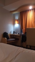 A cozy hotel room featuring a small desk area with a bag on the chair, a bottle of water, and a kettle. An orange curtain hangs beside the desk, softly lit by a nearby wall lamp. The corner of a bed with white sheets is visible in the foreground.