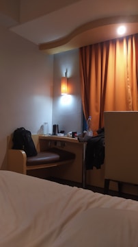 A cozy hotel room featuring a small desk area with a bag on the chair, a bottle of water, and a kettle. An orange curtain hangs beside the desk, softly lit by a nearby wall lamp. The corner of a bed with white sheets is visible in the foreground.