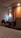 A cozy hotel room featuring a small desk area with a bag on the chair, a bottle of water, and a kettle. An orange curtain hangs beside the desk, softly lit by a nearby wall lamp. The corner of a bed with white sheets is visible in the foreground.