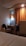 A cozy hotel room featuring a small desk area with a bag on the chair, a bottle of water, and a kettle. An orange curtain hangs beside the desk, softly lit by a nearby wall lamp. The corner of a bed with white sheets is visible in the foreground.