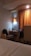 A cozy hotel room featuring a small desk area with a bag on the chair, a bottle of water, and a kettle. An orange curtain hangs beside the desk, softly lit by a nearby wall lamp. The corner of a bed with white sheets is visible in the foreground.