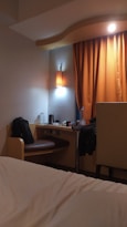 A cozy hotel room featuring a small desk area with a bag on the chair, a bottle of water, and a kettle. An orange curtain hangs beside the desk, softly lit by a nearby wall lamp. The corner of a bed with white sheets is visible in the foreground.