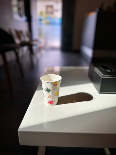 A paper cup adorned with colorful designs, such as squares and stars, sits on a white table. Sunlight casts a shadow of the cup onto the table. Chairs and a blurred background are visible, suggesting an indoor setting with natural light streaming in.