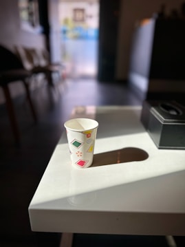 A paper cup adorned with colorful designs, such as squares and stars, sits on a white table. Sunlight casts a shadow of the cup onto the table. Chairs and a blurred background are visible, suggesting an indoor setting with natural light streaming in.
