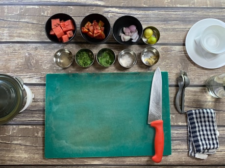 A kitchen setup with a wooden table surface, including a green cutting board, a chef's knife with a red handle, and various cooking ingredients in bowls such as watermelon cubes, chopped tomatoes, shallots, lime, spices, and herbs. A metal citrus squeezer, a striped kitchen towel, and a glass jar are also visible.