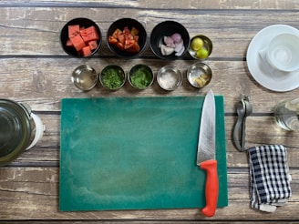 A kitchen setup with a wooden table surface, including a green cutting board, a chef's knife with a red handle, and various cooking ingredients in bowls such as watermelon cubes, chopped tomatoes, shallots, lime, spices, and herbs. A metal citrus squeezer, a striped kitchen towel, and a glass jar are also visible.