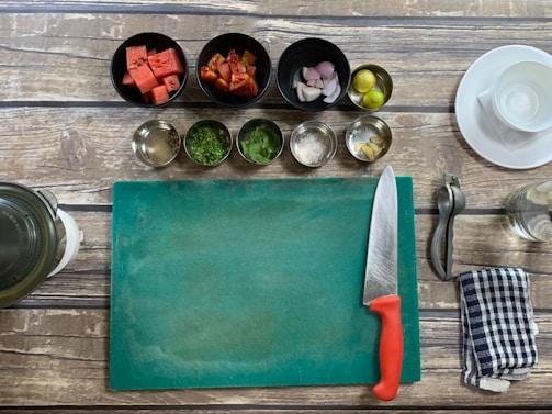 A kitchen setup with a wooden table surface, including a green cutting board, a chef's knife with a red handle, and various cooking ingredients in bowls such as watermelon cubes, chopped tomatoes, shallots, lime, spices, and herbs. A metal citrus squeezer, a striped kitchen towel, and a glass jar are also visible.