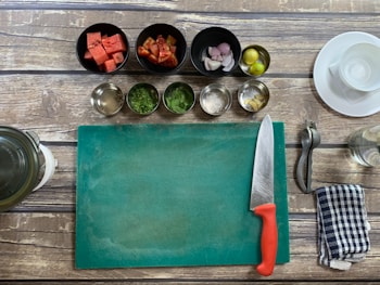 A kitchen setup with a wooden table surface, including a green cutting board, a chef's knife with a red handle, and various cooking ingredients in bowls such as watermelon cubes, chopped tomatoes, shallots, lime, spices, and herbs. A metal citrus squeezer, a striped kitchen towel, and a glass jar are also visible.