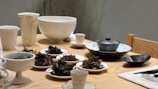A collection of ceramic bowls and plates arranged on a wooden table, featuring various dried tea leaves and small servings of food. The scene includes cups, a teapot, and other dishware, creating a serene and orderly setting.