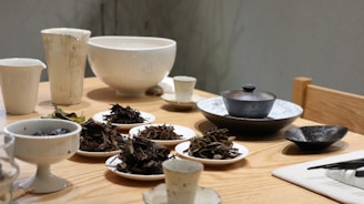 A collection of ceramic bowls and plates arranged on a wooden table, featuring various dried tea leaves and small servings of food. The scene includes cups, a teapot, and other dishware, creating a serene and orderly setting.