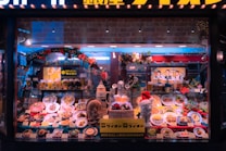 A window display showcasing a variety of plated dishes including pasta, steaks, and desserts, as well as drinks like beer in glasses. There is a festive decoration with flowers and a Santa Claus figurine wearing a red hat. The background features a large image of people holding beer mugs and there are bright, warm lights illuminating the scene.