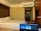 A hotel room with a neatly made bed, featuring multiple pillows. A laptop with its screen open is placed in the foreground, partially visible. The room's door is in the background, and the walls are painted a neutral color. A television is mounted beside the wall near the wooden panels.