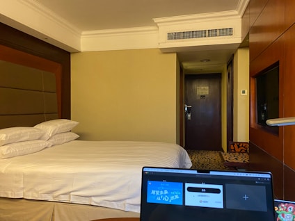 A hotel room with a neatly made bed, featuring multiple pillows. A laptop with its screen open is placed in the foreground, partially visible. The room's door is in the background, and the walls are painted a neutral color. A television is mounted beside the wall near the wooden panels.