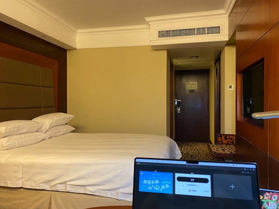 A hotel room with a neatly made bed, featuring multiple pillows. A laptop with its screen open is placed in the foreground, partially visible. The room's door is in the background, and the walls are painted a neutral color. A television is mounted beside the wall near the wooden panels.
