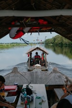 A wooden boat is traveling along a calm river, surrounded by lush green vegetation. There are several people on the boat, with life vests and a lifebuoy visible. The interior of the boat features tables with drinks and food items.
