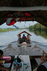 A wooden boat is traveling along a calm river, surrounded by lush green vegetation. There are several people on the boat, with life vests and a lifebuoy visible. The interior of the boat features tables with drinks and food items.