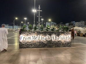 At night, a well-lit sign made of stone with the words 'From Medina To The World' is displayed prominently. Behind the sign, various green plants are placed, and in the background, a cityscape with modern streetlights, buildings, and a few people are visible. A person dressed in traditional attire is walking near the sign.