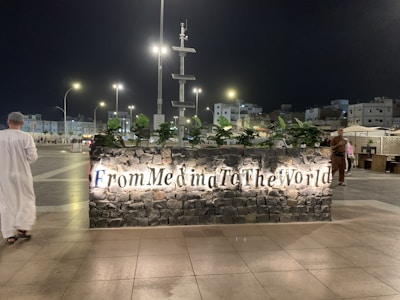 At night, a well-lit sign made of stone with the words 'From Medina To The World' is displayed prominently. Behind the sign, various green plants are placed, and in the background, a cityscape with modern streetlights, buildings, and a few people are visible. A person dressed in traditional attire is walking near the sign.