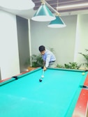 A person is playing pool on a bright blue table. The player is focused on aiming a shot using a cue stick. Overhead, there are green pendant lights providing illumination. The room has a modern look with potted plants surrounding the area.