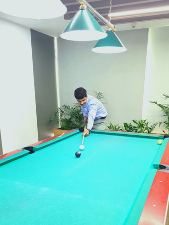 A person is playing pool on a bright blue table. The player is focused on aiming a shot using a cue stick. Overhead, there are green pendant lights providing illumination. The room has a modern look with potted plants surrounding the area.