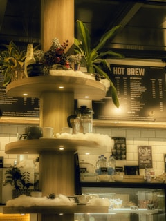 A cozy coffee shop interior featuring a wooden shelf with festive decorations such as pine cones, a small plant, and a few wrapped presents. The background showcases a menu board labeled 'Hot Brew', and various items like coffee cups, mugs, and a coffee press. Soft lighting and warm tones give the scene a comforting ambiance.