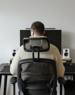 A person is seated in an ergonomic chair facing a desk. The desk features a large monitor, two speakers, and a keyboard with a neutral background.