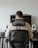 A person is seated in an ergonomic chair facing a desk. The desk features a large monitor, two speakers, and a keyboard with a neutral background.