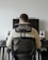 A person is seated in an ergonomic chair facing a desk. The desk features a large monitor, two speakers, and a keyboard with a neutral background.
