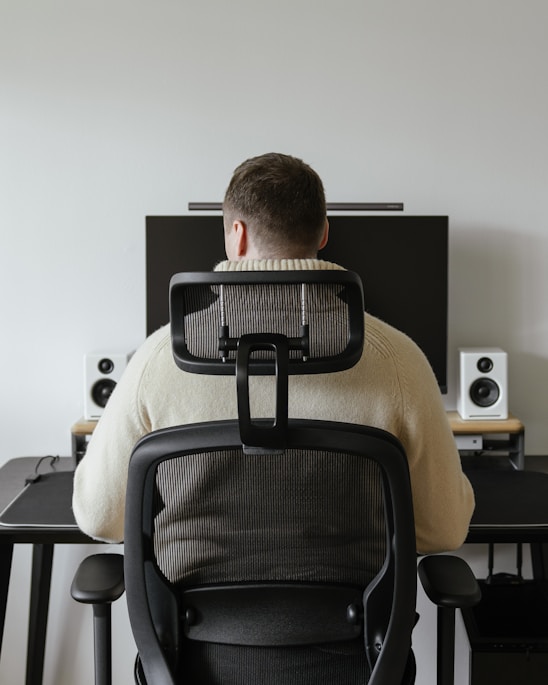 A person is seated in an ergonomic chair facing a desk. The desk features a large monitor, two speakers, and a keyboard with a neutral background.