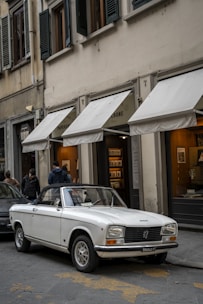 A classic white convertible car is parked on a cobblestone street in front of a traditional building with multiple awnings covering the shop entrances. Two people are walking on the sidewalk, and large windows with green shutters are located on the building's upper levels.