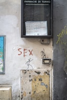 A wall with graffiti featuring stick figures and the word 'SEX' painted in red. Above the graffiti, there is a display case labeled 'FARMACIE DI TURNO' containing a notice or schedule. The wall is worn and peeling, with exposed plaster and a patched-up section.