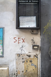 A wall with graffiti featuring stick figures and the word 'SEX' painted in red. Above the graffiti, there is a display case labeled 'FARMACIE DI TURNO' containing a notice or schedule. The wall is worn and peeling, with exposed plaster and a patched-up section.