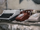 A collection of vintage communication devices, including a typewriter and two rotary dial telephones, is placed on a rough stone surface. The devices show signs of wear and age, enhancing their nostalgic appearance.