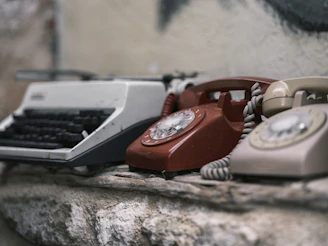 A vintage facsimile machine sitting on a wooden desk with old newspapers and a rotary phone nearby.