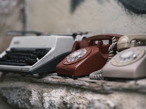 A collection of vintage communication devices, including a typewriter and two rotary dial telephones, is placed on a rough stone surface. The devices show signs of wear and age, enhancing their nostalgic appearance.