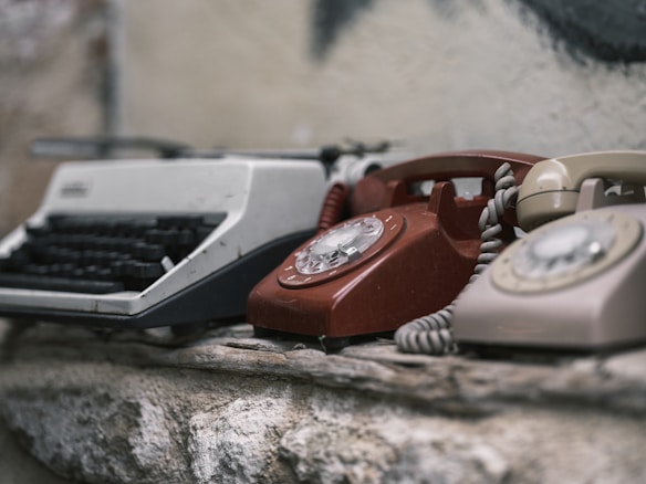 A collection of vintage communication devices, including a typewriter and two rotary dial telephones, is placed on a rough stone surface. The devices show signs of wear and age, enhancing their nostalgic appearance.