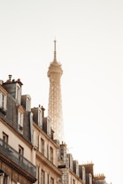 The image features the upper section of the Eiffel Tower soaring into a clear sky, with the warm glow of the morning or evening sun reflecting off its structure. In the foreground, elegant Parisian buildings with classic rooftops and windows line the street, creating a harmonious blend of architecture.