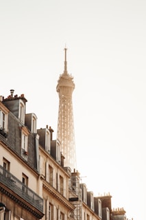 The image features the upper section of the Eiffel Tower soaring into a clear sky, with the warm glow of the morning or evening sun reflecting off its structure. In the foreground, elegant Parisian buildings with classic rooftops and windows line the street, creating a harmonious blend of architecture.