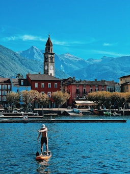 A picturesque lakeside scene featuring a person paddleboarding on a calm body of water. In the background, there are colorful buildings with a prominent clock tower rising above them. The distant mountains create a scenic backdrop under a clear blue sky.